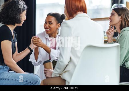 sorridente donna afro-americana che si tiene per mano con una ragazza multirazziale vicino a una rossa motivazione coach seduta con un caffè da asporto su un foregro sfocato Foto Stock