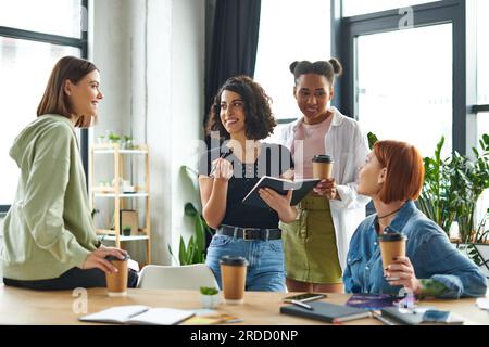 donna ottimista multirazziale che tiene penna e taccuino durante la comunicazione con un gruppo eterogeneo di amiche multietniche in donne interessano cl Foto Stock