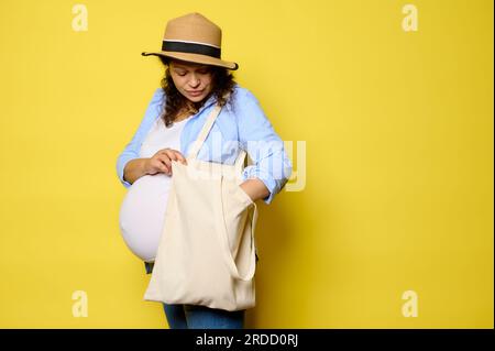 Autentico ritratto di donna incinta latino-americana dai capelli ricci con shopper di lino, borsa per la spesa ecologica, isolata sullo sfondo giallo dello studio. Sustainabil Foto Stock