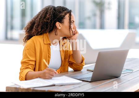 Noiosa donna dai capelli ricci brasiliana o latinoamericana, freelance, studentessa, seduta all'aperto, vicino al campus, con un notebook, sembra triste di lato, è esausto, annoiato e premuroso Foto Stock