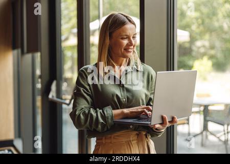 Donna d'affari sorridente che lavora su un computer portatile in un accogliente ufficio in piedi vicino alle finestre Foto Stock