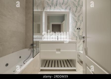 WC decorato in un design moderno con vasca idromassaggio, armadio in legno bianco con cassetti e specchio quadrato con cornice in legno coordinata Foto Stock