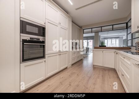 Una spaziosa cucina con pareti ricoperte di armadi di legno bianco da pavimento a soffitto con pavimenti di legno chiaro Foto Stock