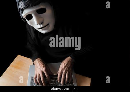 Hacker vestito di nero con maschera e cappuccio che scrivono su un computer mentre guardano il concetto di cybercrime Foto Stock