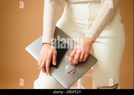 Primo piano di una donna che tiene uno smartphone, un computer portatile e degli occhiali sulla gamba Foto Stock