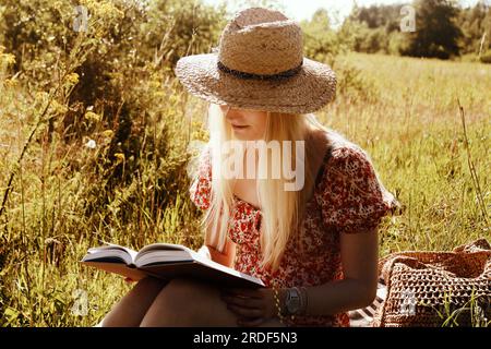 Giovane donna bionda con un cappello di paglia e un libro in estate Foto Stock