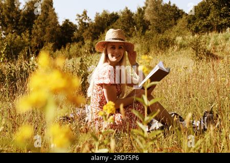 Giovane donna bionda con un cappello di paglia e un libro in estate Foto Stock