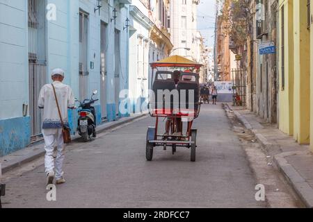 L'Avana, Cuba - 27 maggio 2023: Un uomo afro-caraibico che indossa abiti bianchi cammina in una strada cittadina. Un bicitaxi o pedicab guida nel centro residenziale Foto Stock