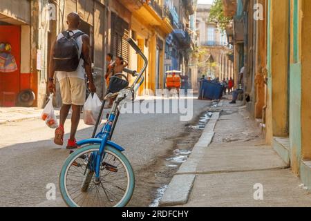 L'Avana, Cuba - 27 maggio 2023: Un uomo afro-caraibico cammina con due borse di generi alimentari in una strada cittadina. Una bicicletta è ferma accanto al marciapiede. Lifest Foto Stock