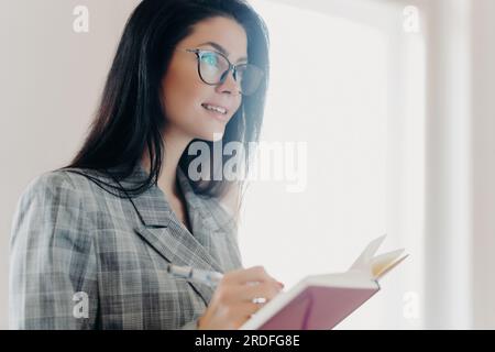 Una donna europea bruna premurosa in abiti formali e occhiali trasparenti scrive le informazioni testuali in blocchi note, in una concentrazione profonda in ambienti interni. Foto Stock