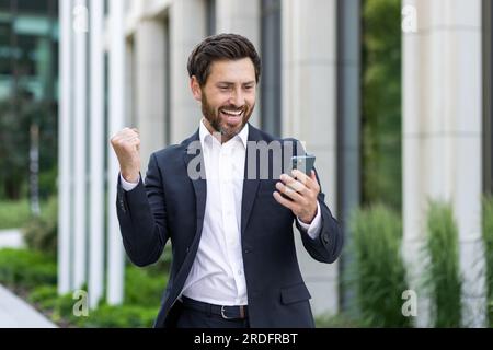 Uomo d'affari maturo ed esperto con telefono che celebra il successo trionfo della vittoria, il capo in tuta aziendale ha ricevuto una buona notifica online, uomo fuori dall'edificio degli uffici che tiene la mano sul gesto del vincitore. Foto Stock