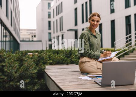 Analista donna che lavora con documenti e utilizza un notebook seduto all'esterno su un edificio moderno Foto Stock