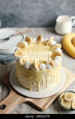 Torta di banana e noci con ganache di cioccolato bianco sulla parte superiore, panna montata e fette di banana. Tagliere in legno e sfondo blu. Foto Stock