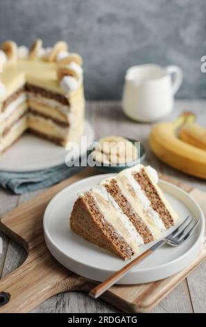 Torta di banana e noci con ganache di cioccolato bianco sulla parte superiore, panna montata e fette di banana. Tagliere in legno e sfondo blu. Foto Stock