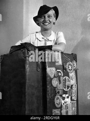 New York, New York: c. 1933 Una donna si trova con il suo baule a vapore che è coperto da cartelli per hotel e viaggi Foto Stock