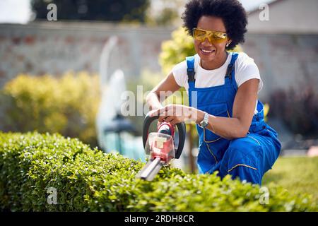 Allegra giovane donna afro-americana che utilizza tagliasiepi elettricisti per tagliare siepi all'aperto nel cortile, lavorando in giardino. Stile di vita, casa, all'aperto Foto Stock