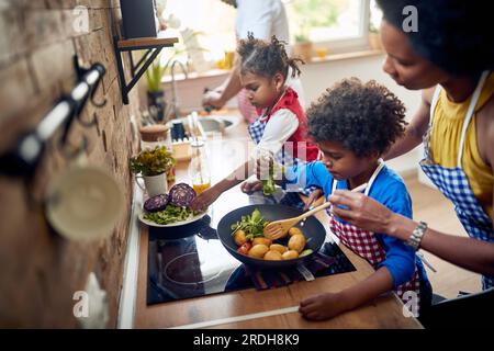 In cucina, mentre una madre prepara sapientemente un pasto delizioso, mentre i suoi figli, un ragazzo e una ragazza, si uniscono con ansia al processo di cottura. Dietro Foto Stock