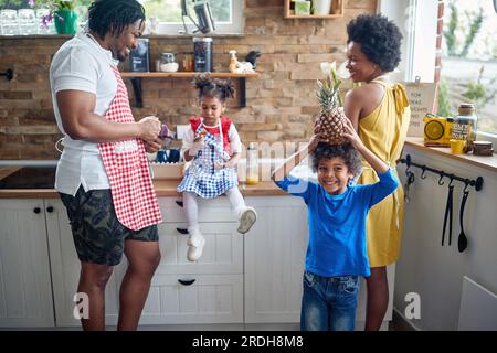 Bella e gioiosa famiglia in una cucina domestica insieme, bambino che tiene l'ananas sulla testa come una corona. Concetto di casa, famiglia, stile di vita. Foto Stock