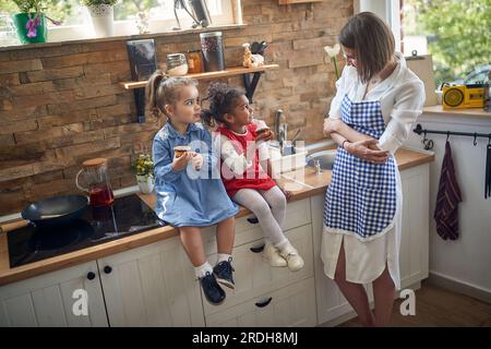 Due ragazze, sedute insieme in cucina, assaporando deliziosi muffin. In piedi accanto a loro, la loro madre travolge con orgoglio e gioia, il suo sorriso a testa Foto Stock