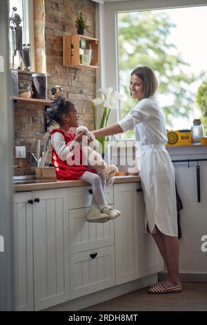 La bambina si sta godendo un delizioso muffin in in una mano mentre tiene il suo amato orsacchiotto nell'altra. Al suo fianco si trova la sua premurosa madre, dili Foto Stock