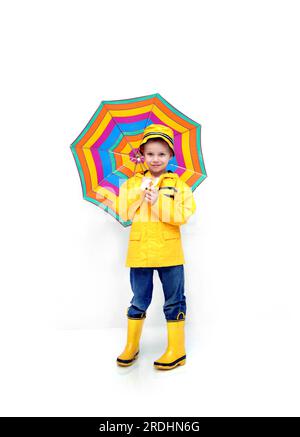 Little boy stands prepared for rain.  He is wearing a yellow raincoat and hat.  He is carrying a striped umbrella and has his jeans tucked into yellow Stock Photo