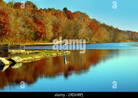 Il pescatore solitario getta la sua linea nel piccolo lago nel nord dell'Arkansas. La luce del mattino presto riflette il blu nella superficie cristallina del lago e il fogliame autunnale aggiunge Foto Stock