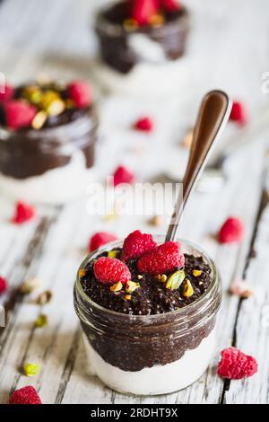 Budino di chia al cioccolato fondente con lamponi e pistacchi in cima in un vaso di vetro su sfondo di legno bianco. Servita con panna montata al cocco. Foto Stock