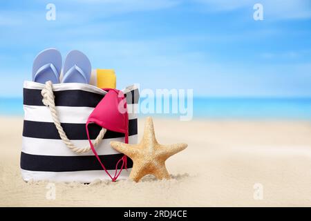 Borsa con zona bikini e accessori sulla soleggiata spiaggia dell'oceano, spazio per messaggi. Vacanze estive Foto Stock