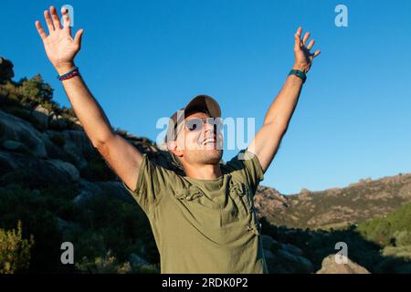 Uomo vestito con t-shirt verde militare, berretto e occhiali da sole che si gode una giornata in montagna Foto Stock