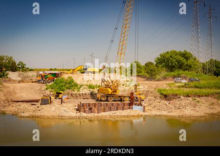 Oltre l'acqua è presente una gru mobile gialla obsoleta con martello pneumatico. Pali di metallo installati lungo la riva del fiume integrati per la fondazione di ponti presso il c Foto Stock