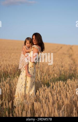 Una giovane bella signora che tiene la sua figlia in un campo al tramonto. Una donna con un bambino in un bellissimo vestito giallo posa al tramonto in un campo. Foto Stock