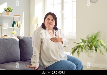 Donna in sovrappeso con malattie cardiache seduto sul divano e sofferente di dolore al petto Foto Stock