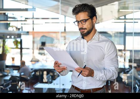 Un giovane uomo d'affari latino serio che controlla i documenti finanziari in ufficio. Foto Stock