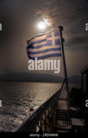 Bandiera greca che sventola davanti al sole con cielo parzialmente nuvoloso. Bandiera della Grecia retroilluminata su un traghetto. Foto Stock