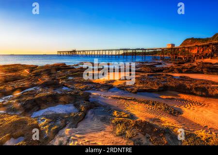 Lungo e storico molo presso la spiaggia Middle Camp di Catherine Hill Bay, cittadina costiera australiana, dalla scenografica spiaggia di sabbia con bassa marea. Foto Stock