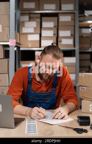 Dipendente maschio che pianifica la distribuzione dei prodotti nello spazio di magazzino, scrivendo l'indirizzo sull'adesivo presso il magazzino di distribuzione Foto Stock