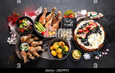 Cena con pollo, verdure, torta, insalata e frutta su sfondo scuro. Concetto di cibo estivo. Vista dall'alto. Foto Stock