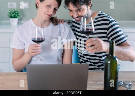 Concetto di festa virtuale. Coppia sorridente seduta al tavolo e videochiamata online con amici o familiari utilizzando il laptop, bevendo vino e toasti Foto Stock
