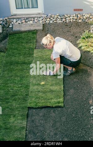 Una donna lavora in un piccolo giardino, posando rotoli di erba nuova su terreno preparato Foto Stock