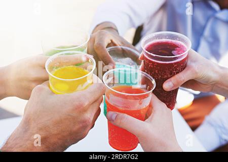 Dettagli sulle mani di un gruppo di giovani che tengono colorate bevande gassate a base di frutta in tazze di plastica, con la luce del sole che filtra attraverso. Foto Stock