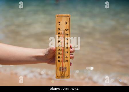 Termometro in mano su uno sfondo spiaggia che mostra temperature elevate. Clima caldo e clima mutevole Foto Stock