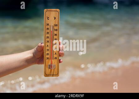 Termometro in mano su uno sfondo spiaggia che mostra temperature elevate. Clima caldo e clima mutevole Foto Stock