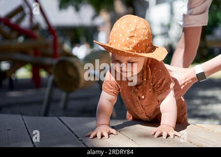 Mamma aiuta il bambino a gattonare. Carino bambino a panama. Ritratti di una bella bambina di 10 mesi. Passeggiate nel parco giochi, in una soleggiata giornata estiva. Foto Stock