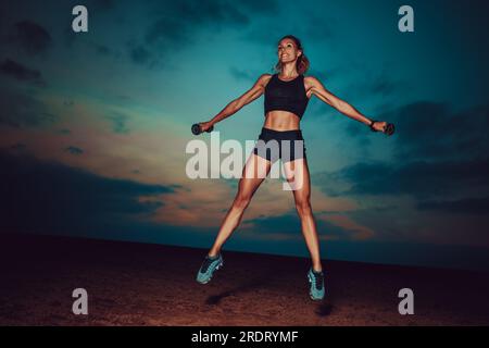 Giovane donna che si forma in forma saltando con i manubri sullo sfondo del cielo al crepuscolo Foto Stock