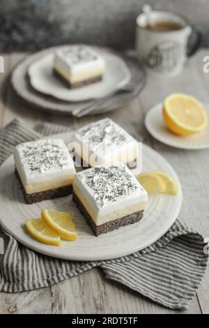 Barrette cremose di limone e semi di papavero su un piatto bianco decorato con fette di limone fresche. Fondo grigio e tovagliolo spelato. Tazza di caffè. Foto Stock