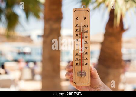 Termometro in mano su uno sfondo spiaggia che mostra temperature elevate. Clima caldo e clima mutevole Foto Stock