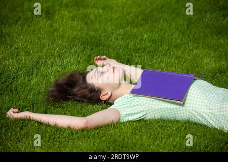 Una giovane donna sdraiata sulla schiena con le braccia tese. Indossa un vestito estivo e ha un libro sul petto. Foto Stock