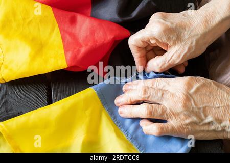 Le mani delle donne cucirono la bandiera della Germania con la bandiera dell'Ucraina sul Consiglio di amministrazione a casa, produzione e lavoro, l'unione di due stati, l'amicizia Foto Stock