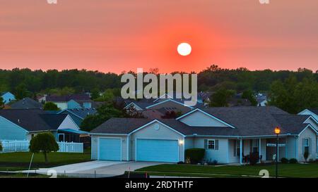 Alba nel cielo rosso-arancione al tramonto sulle case suburbane nello skyline della foresta del quartiere e sull'antenna lampione Foto Stock