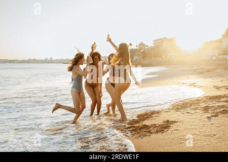 Gioiose giovani donne multirazziali in costume da bagno che ballano, tengono in mano bottiglie di birra, si godono la festa estiva in spiaggia, spazio libero Foto Stock